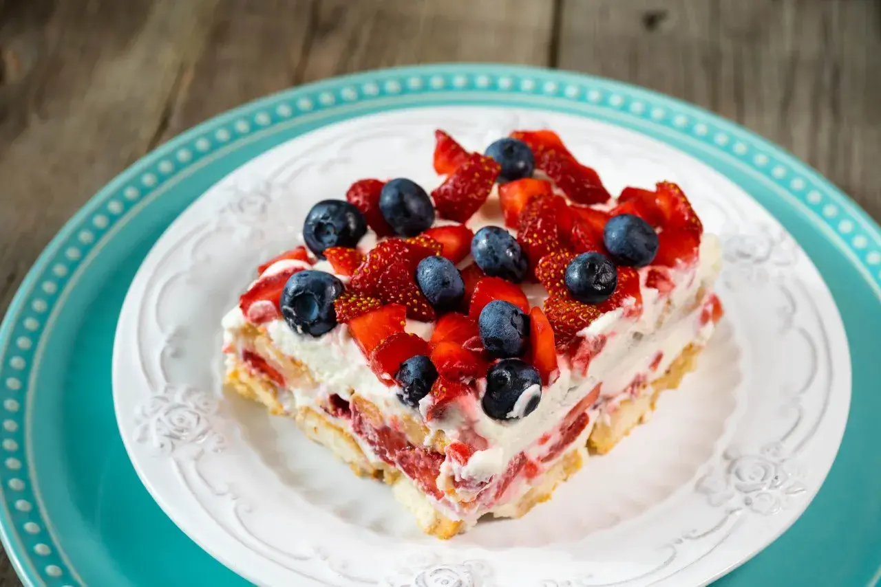 Tranche de tiramisu aux fruits rouges, fraises et myrtilles, sur une assiette blanche ornée, posée sur une assiette turquoise.