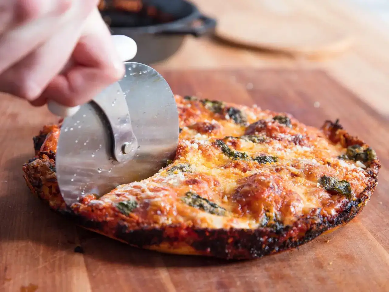 Une main coupe une pizza à la poêle dorée et garnie de fromage fondant et d'épinards sur une planche en bois.