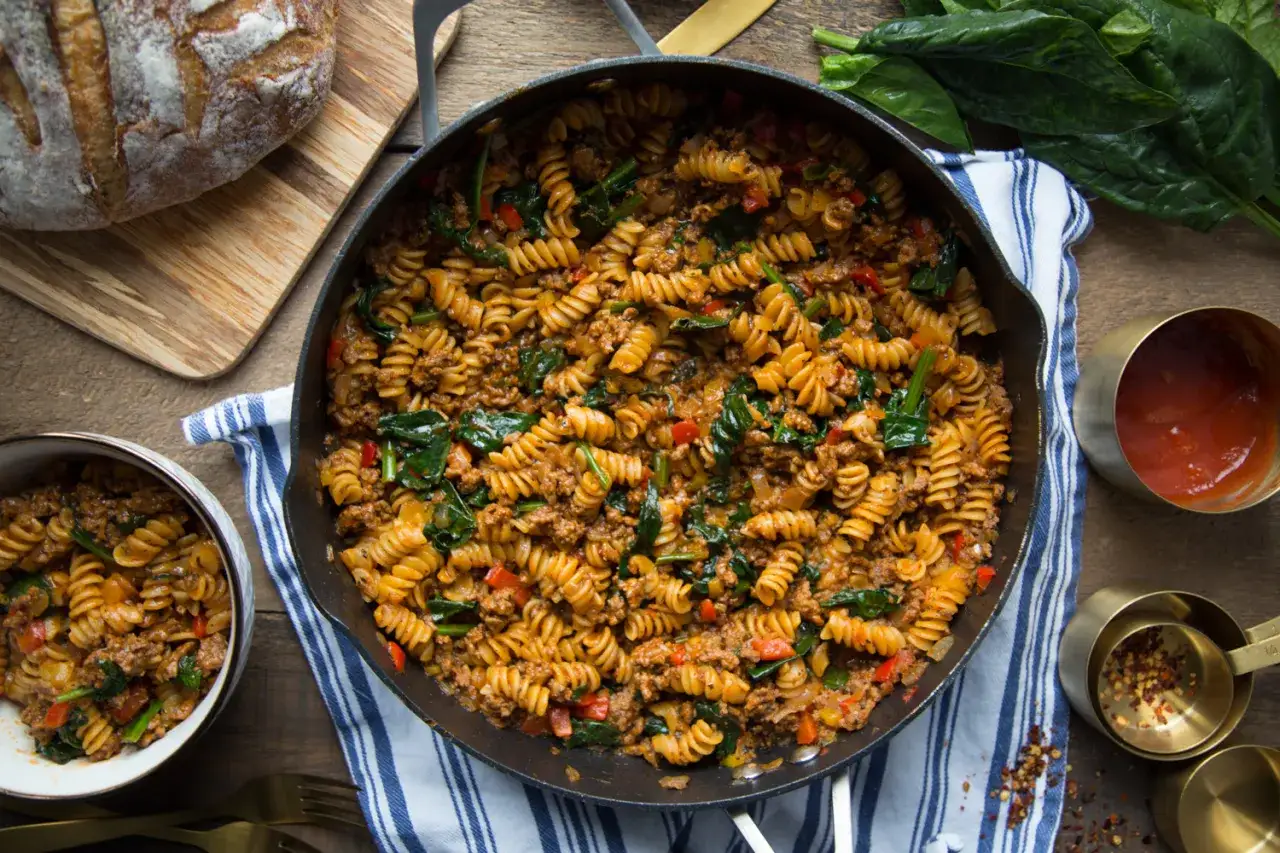 Un plat généreux de pâtes à la viande, avec des fusillis, des épinards et des poivrons, servi dans une poêle en fonte.