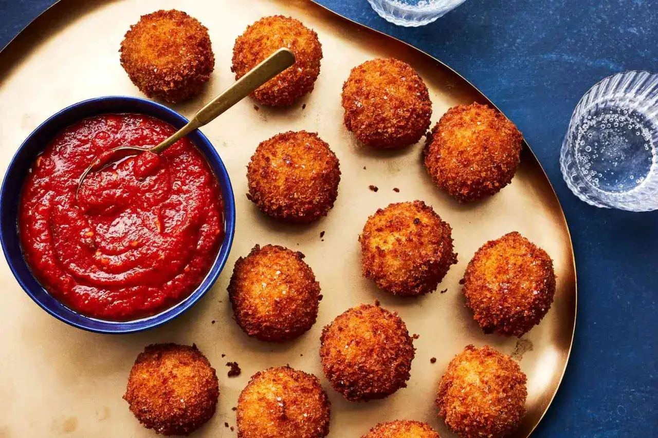 Arancini dorés et croustillants disposés sur un plateau, accompagnés d'une sauce pour arancini rouge vif dans un bol bleu.