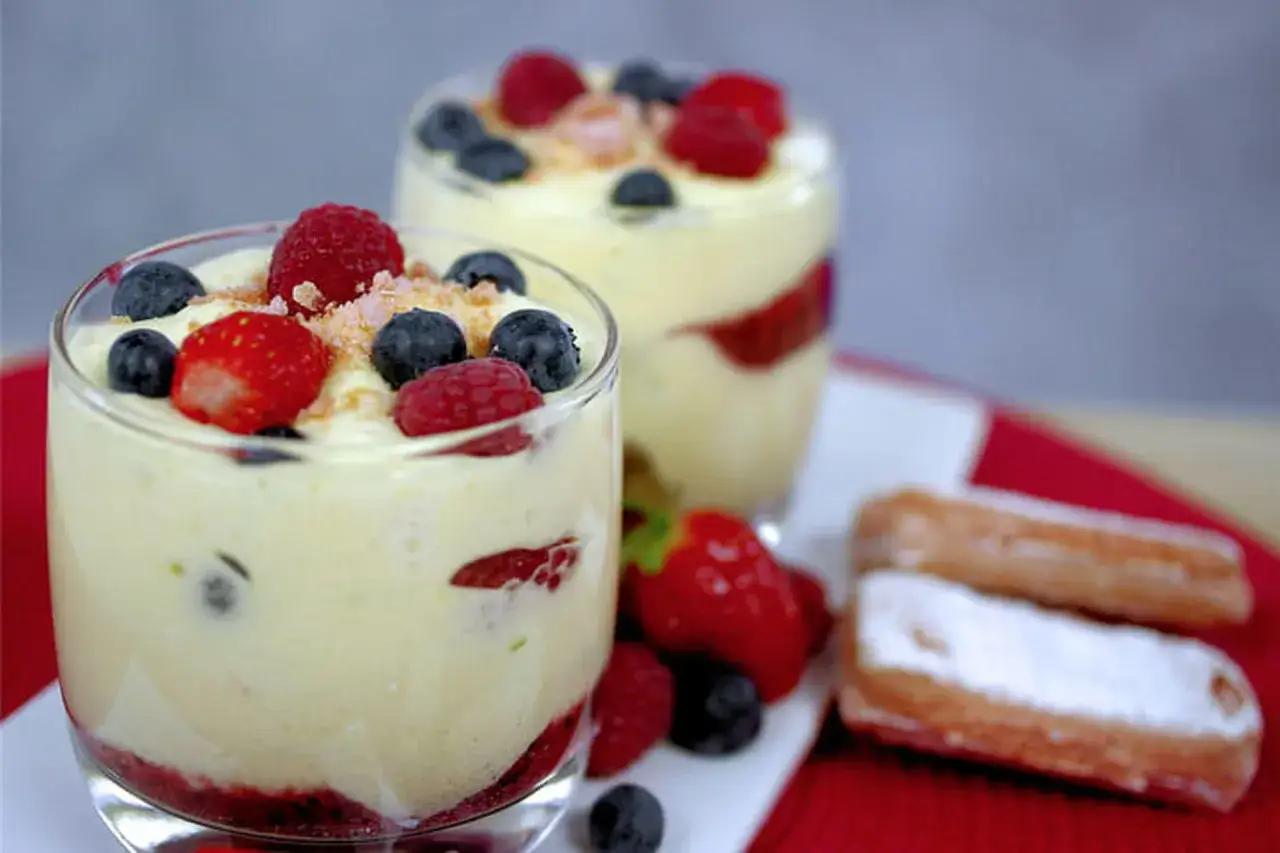 Deux verrines de tiramisu fruitées, garnies de framboises, myrtilles et fraises. Des biscuits à la cuillère saupoudrés de sucre glace sont à côté.