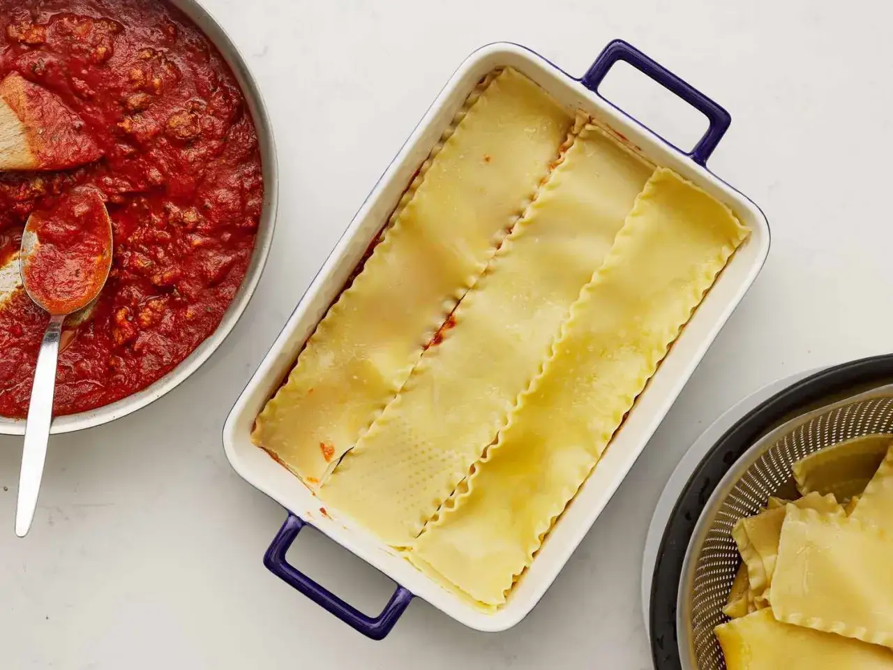 Pr&eacute;paration d'une lasagne maison : sauce tomate dans une po&ecirc;le, p&acirc;tes dans un plat, et p&acirc;tes &eacute;goutt&eacute;es dans une passoire.