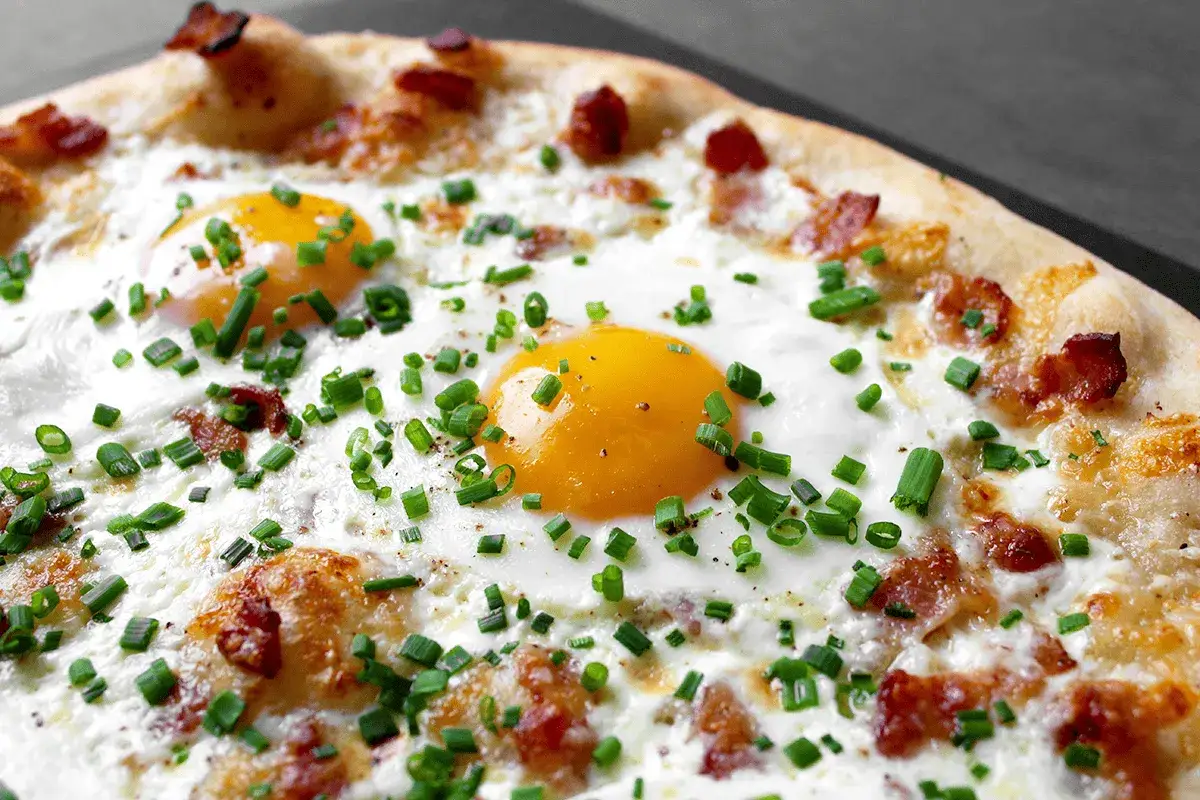Pizza carbonara avec œufs au plat, lardons croustillants et ciboulette fraîche. Un délice pour les yeux et les papilles.