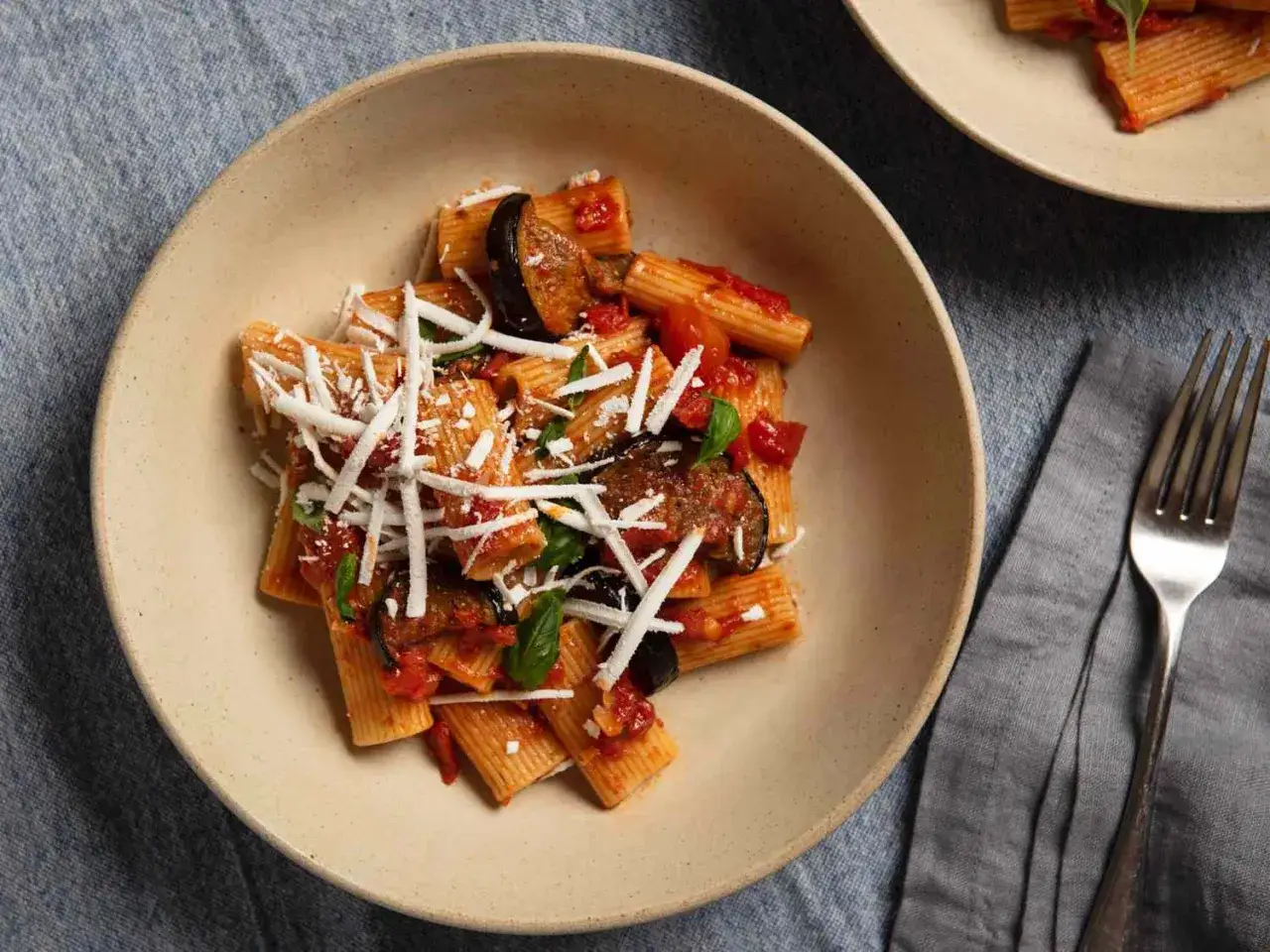 Un plat de pâtes aux aubergines à la sicilienne, garni de fromage râpé et de basilic frais, servi dans un bol en céramique.