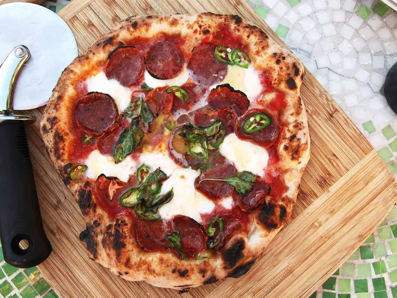 Pizza diavola avec pepperoni, jalapeños et mozzarella fondue, prête à être coupée sur une planche en bois.