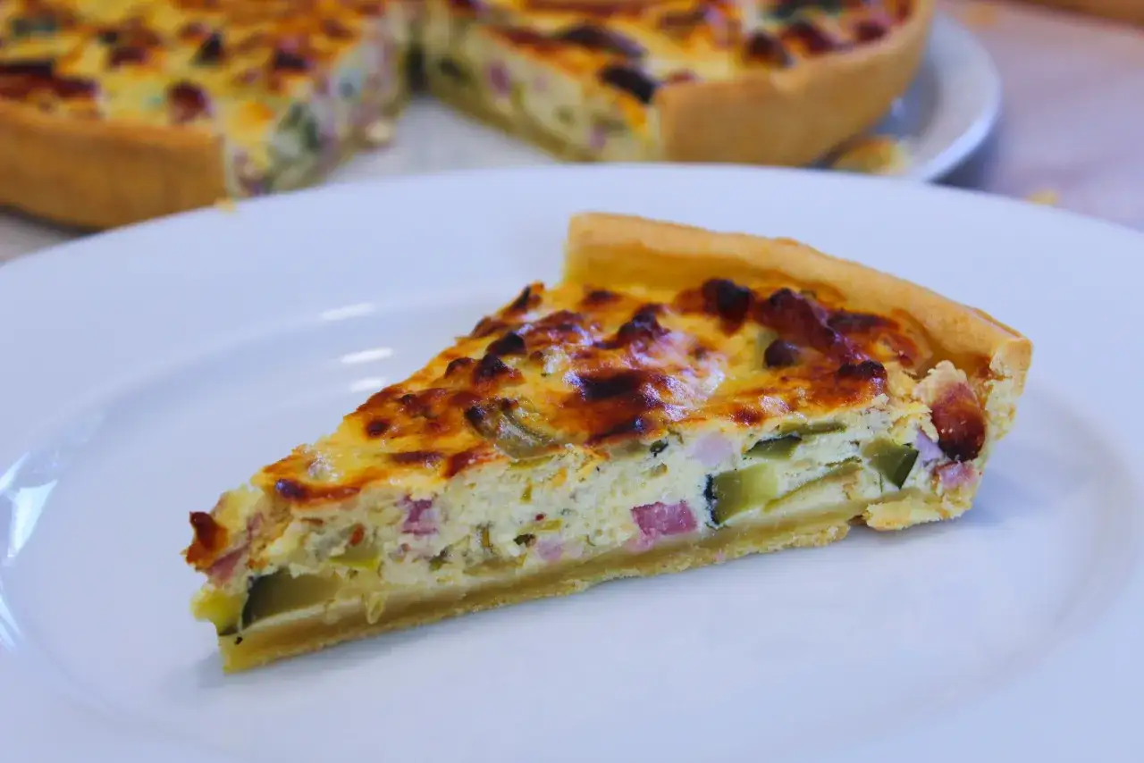Tranche de tarte ricotta courgette, garnie de lardons et de fromage doré, servie sur une assiette blanche.